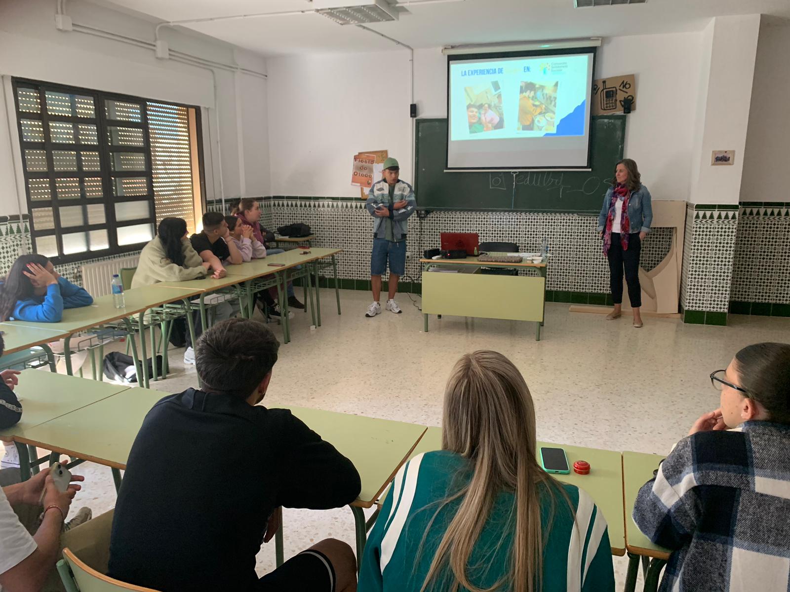 Varias personas sentadas alrededor de mesas en forma de U escuchan a dos personas de pie frente a la clase, junto a una pizarra y un proyector.