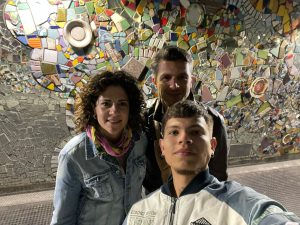 Tres personas posan sonrientes frente a un mural colorido decorado con mosaicos y piezas cerámicas.
