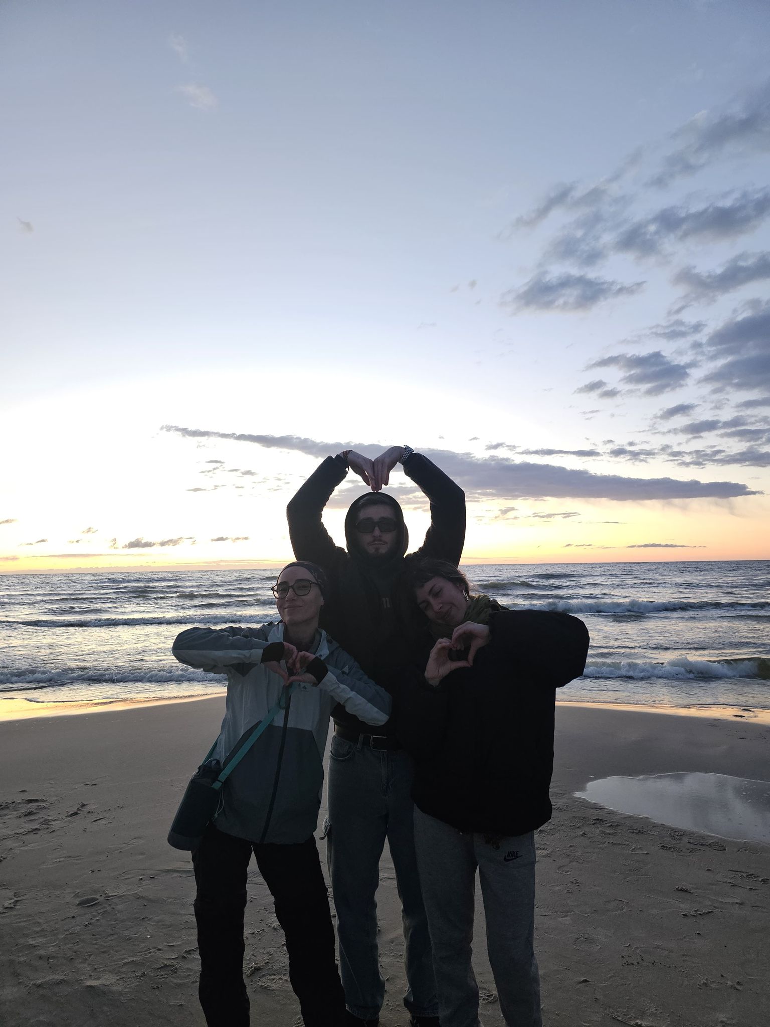 Tres personas posan en la playa al atardecer, haciendo corazones con las manos mientras sonríen frente al mar.