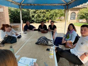 Grupo de personas sentadas alrededor de una mesa al aire libre bajo una carpa, participando en una reunión o actividad formativa.