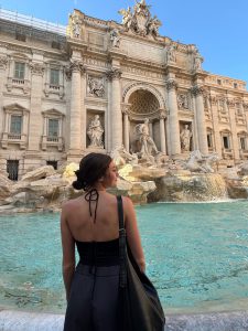 Persona de espaldas contemplando una fuente monumental en una plaza histórica, con esculturas clásicas y cielo despejado.