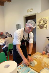 Persona joven con delantal y gorro de cocina cortando masa con un cortador circular en una mesa, durante una actividad culinaria grupal.